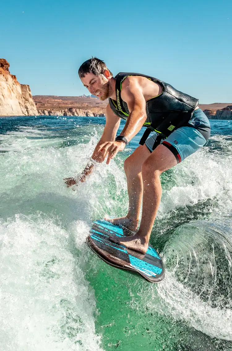 Photo: a wakesurfer in a life vest crouched on the clean green face of a boat wake, hand skimming the water, red canyon shoreline behind.